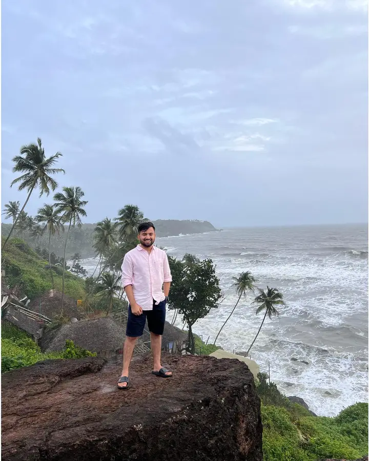 Priyansh standing on a rock above a grey sea, palm trees behind