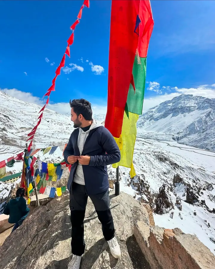 Priyansh on a rock in front of snow-covered mountains, prayer flags overhead